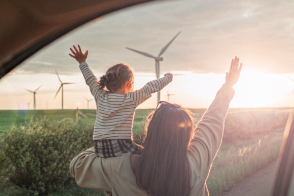 La mère tient sa fille dans ses bras tandis qu'elles lèvent toutes les deux les bras et regardent un champ d'éoliennes.